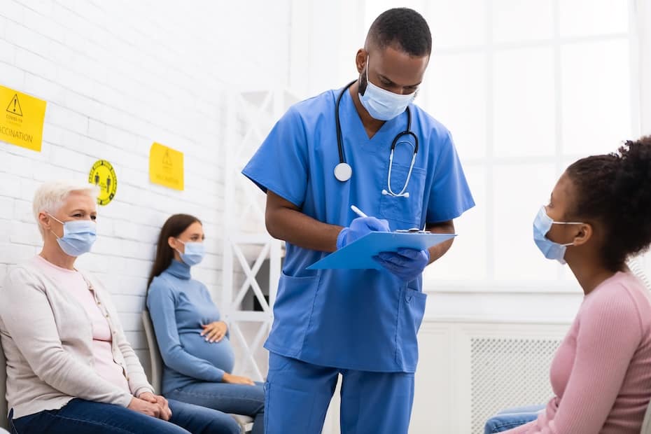 A medical assistant in blue scrubs and a face mask consulting with a patient in a healthcare waiting area.
